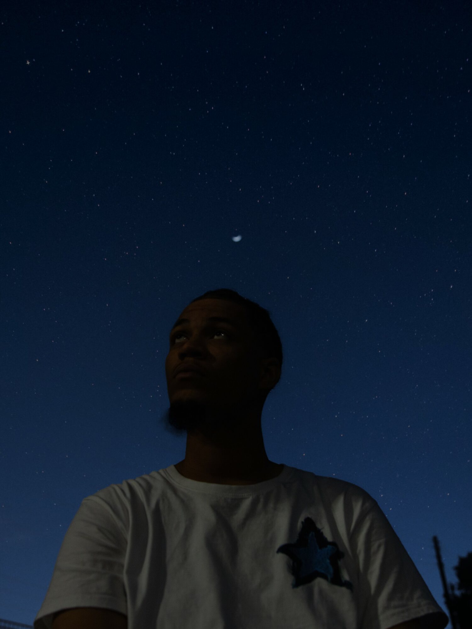 Person with short hair and a white shirt looking up at a starry night sky.