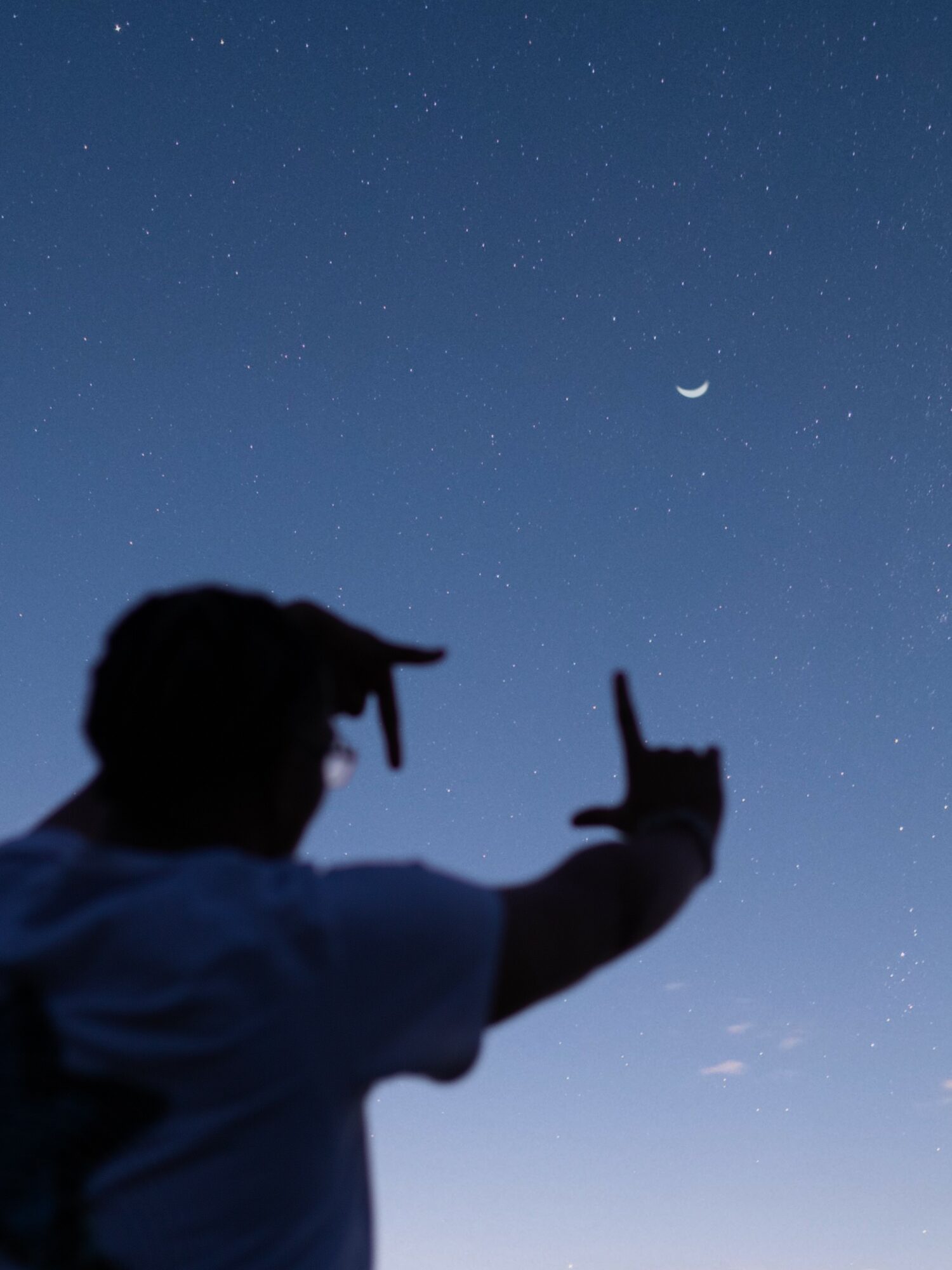 Silhouette of a person pointing at the night sky with a crescent moon and stars visible, background of a clear sky.