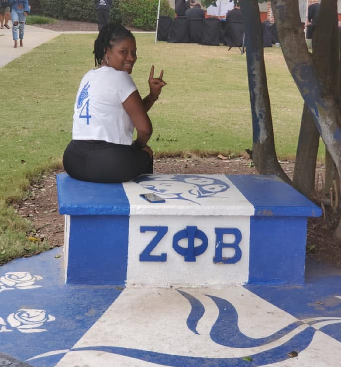 Young woman sitting on a decorated blue and white bench outdoors, making a peace sign with her hand, smiling, with trees and people in background.