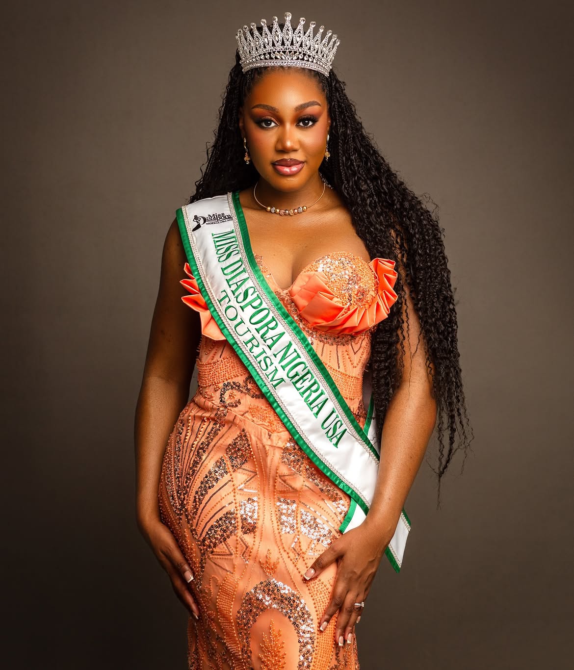 Woman wearing a crown and sash, dressed in an orange gown, standing against a plain background.