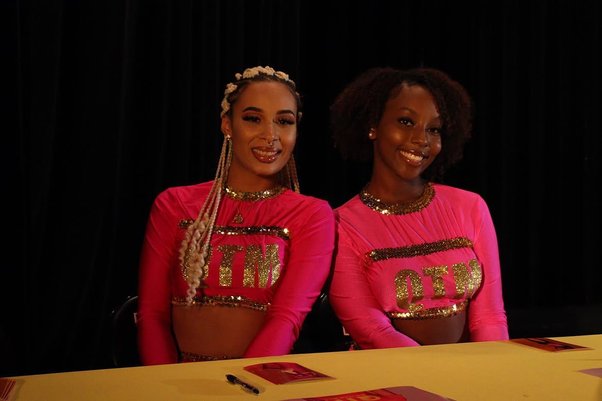 Two young women in pink outfits with gold embellishments, smiling and standing behind a yellow table, black background.
