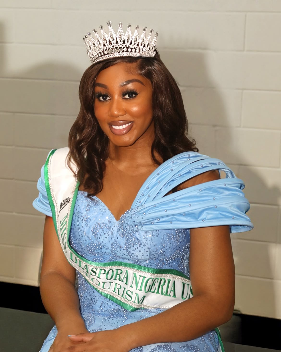 Young woman wearing a crown and sash, smiling, in a blue dress with ruffled sleeves, standing against a plain wall.
