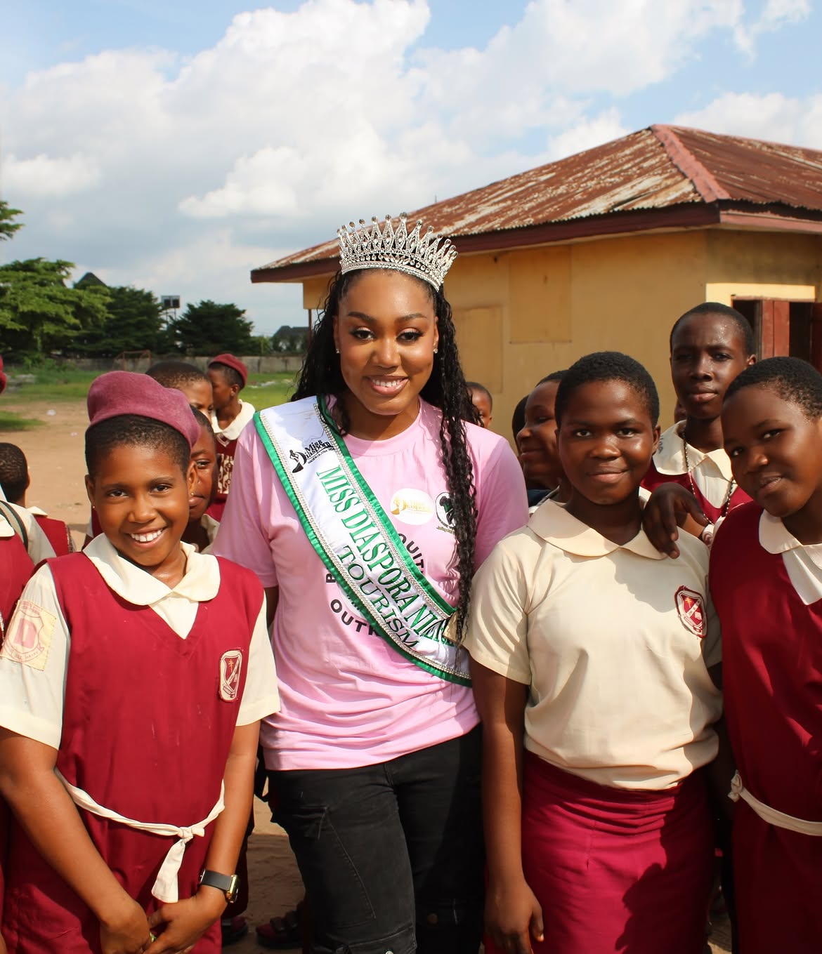 Woman wearing a crown and sash surrounded by children outdoors, smiling, with a building and sky in background.