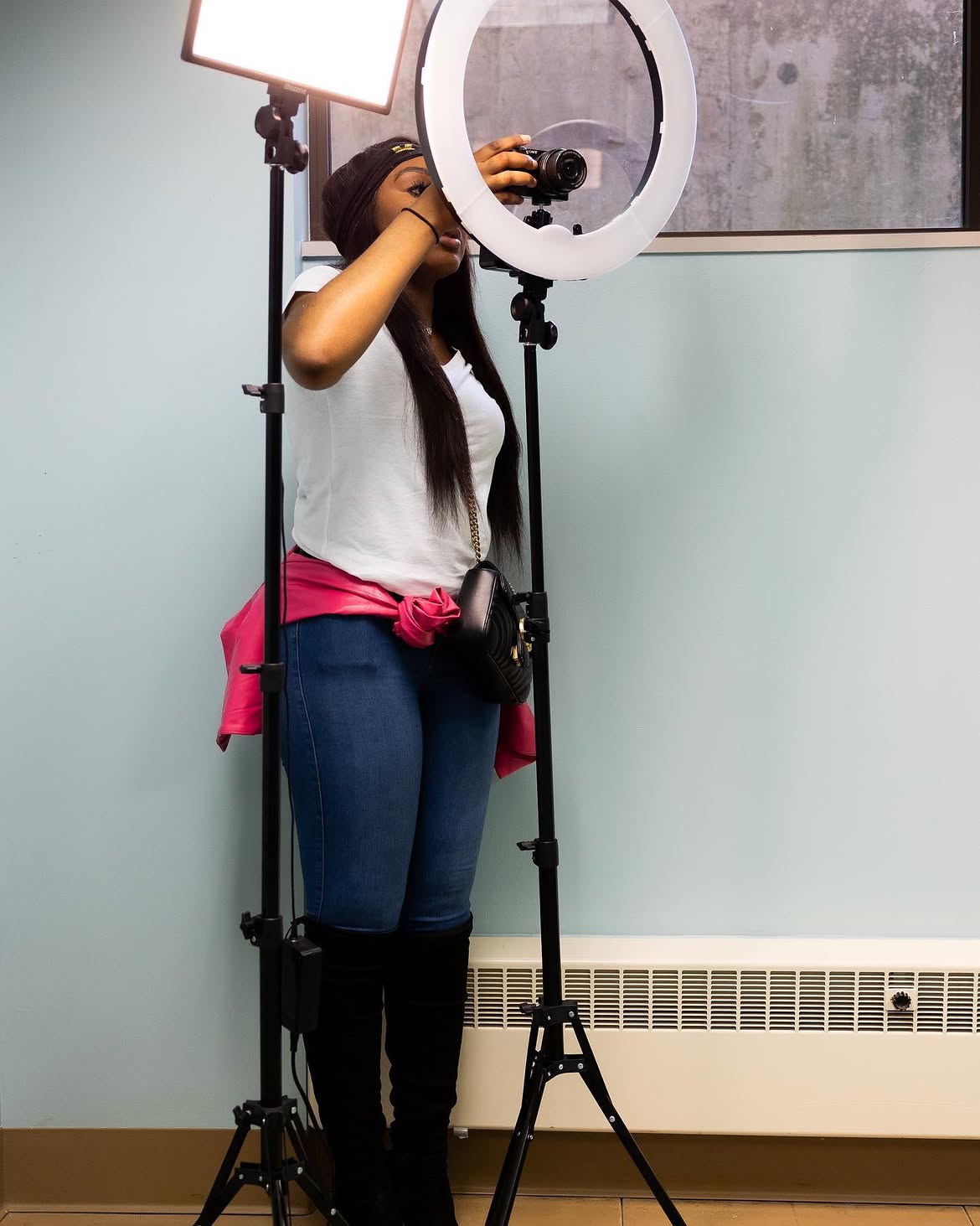 Person using a camera and ring light setup, standing in a room with a light blue wall and radiator.