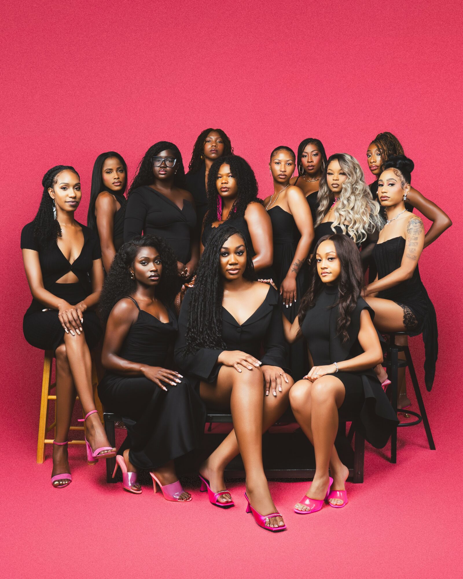 Group of 13 women in black dresses and pink heels against a pink background, some sitting and some standing.