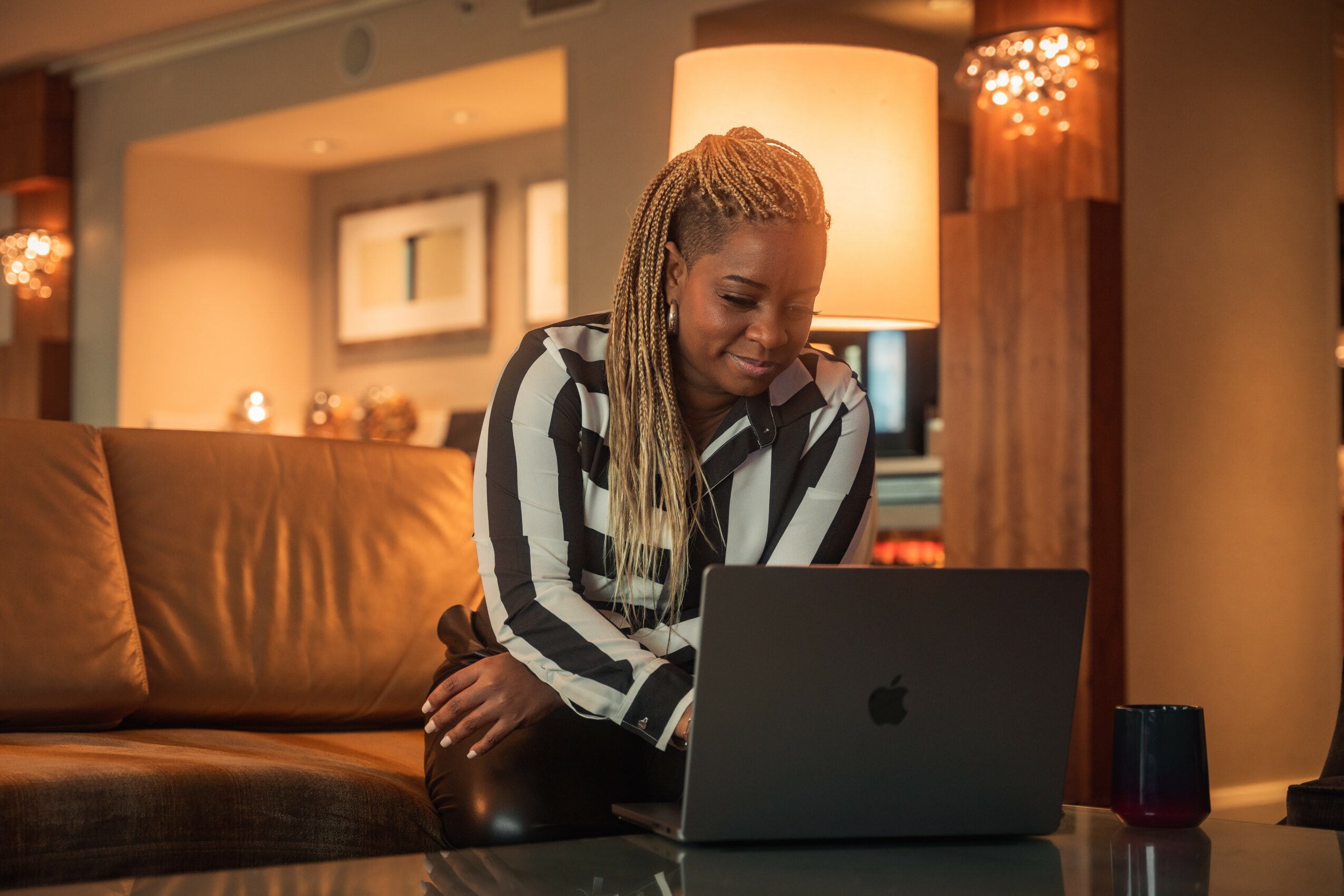 Woman with long braided hair using a laptop on a sofa in a warmly lit living room.