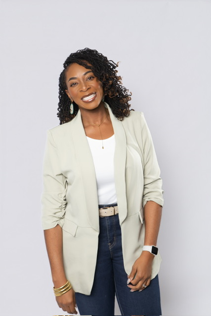 Smiling woman with curly hair wearing a light blazer, white top, and dark jeans, standing against a plain background.