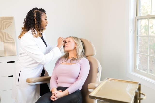 A healthcare professional examines a woman sitting in a medical office, with a window and medical equipment nearby.