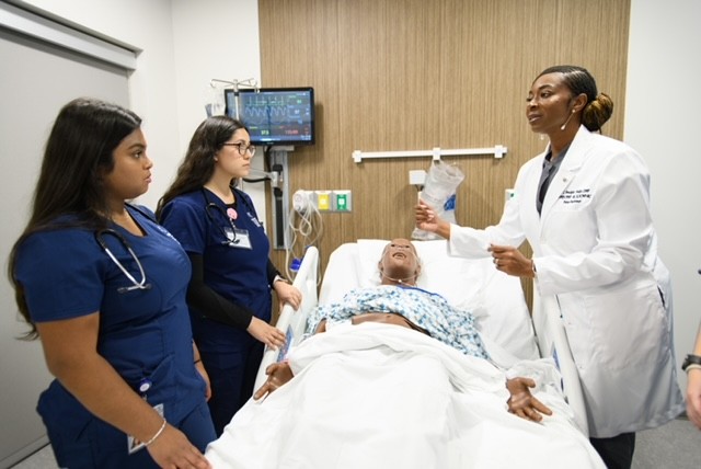Medical professional explains to three nurses around a hospital bed with a patient, in a clinical room with medical equipment.