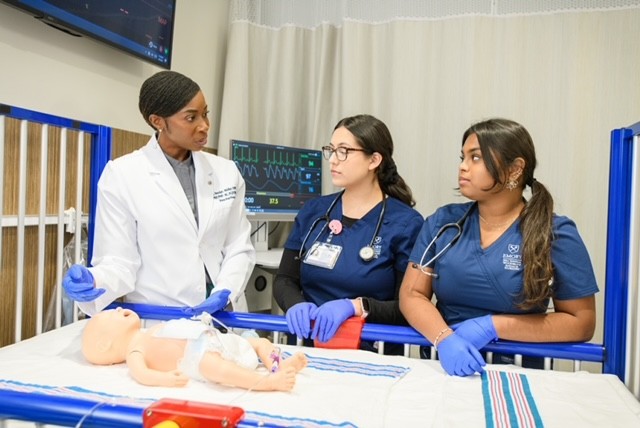 Three healthcare students in scrubs and lab coats discuss a medical mannequin on a hospital bed.
