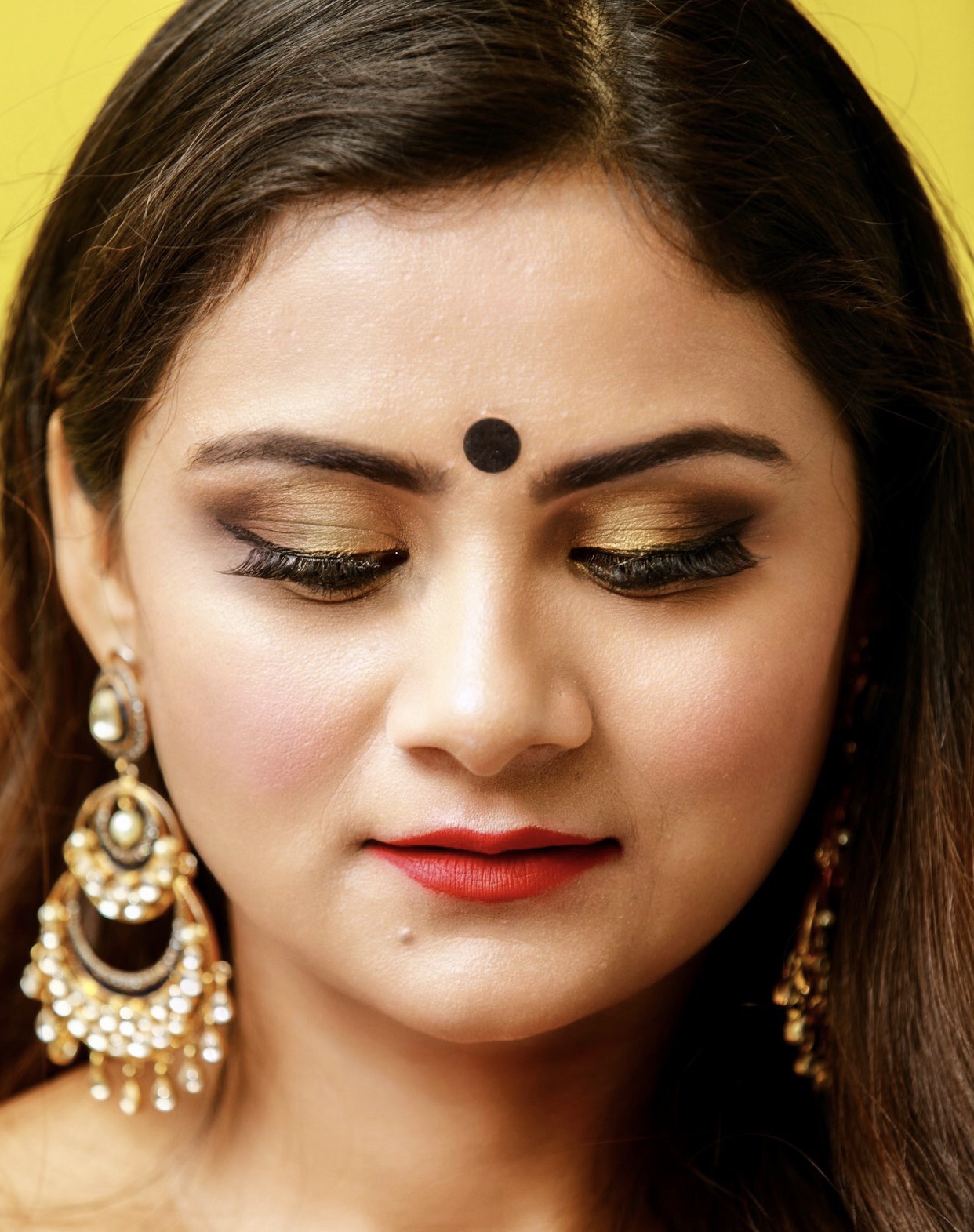 Close-up of a woman with makeup, earrings, and a bindi, looking downward against a yellow background.