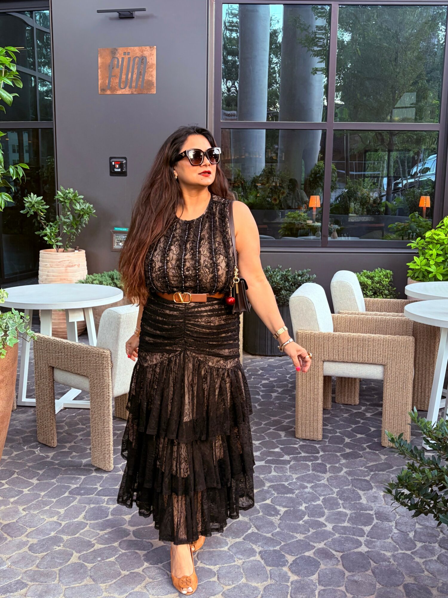 Woman in black dress with sunglasses walking outdoors near tables and chairs, greenery in background.