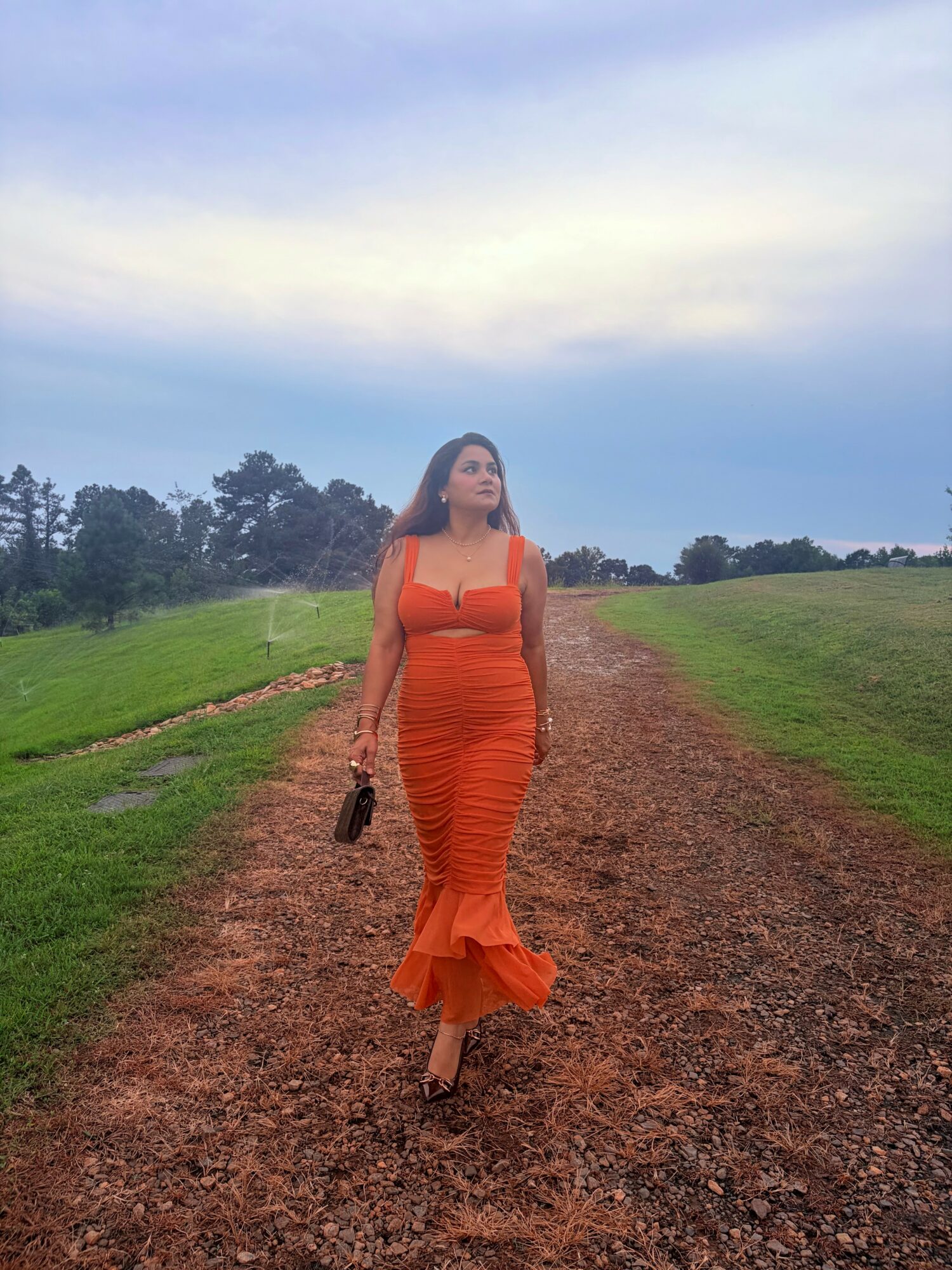 Woman in orange dress walking on a dirt path in a grassy field under a cloudy sky.