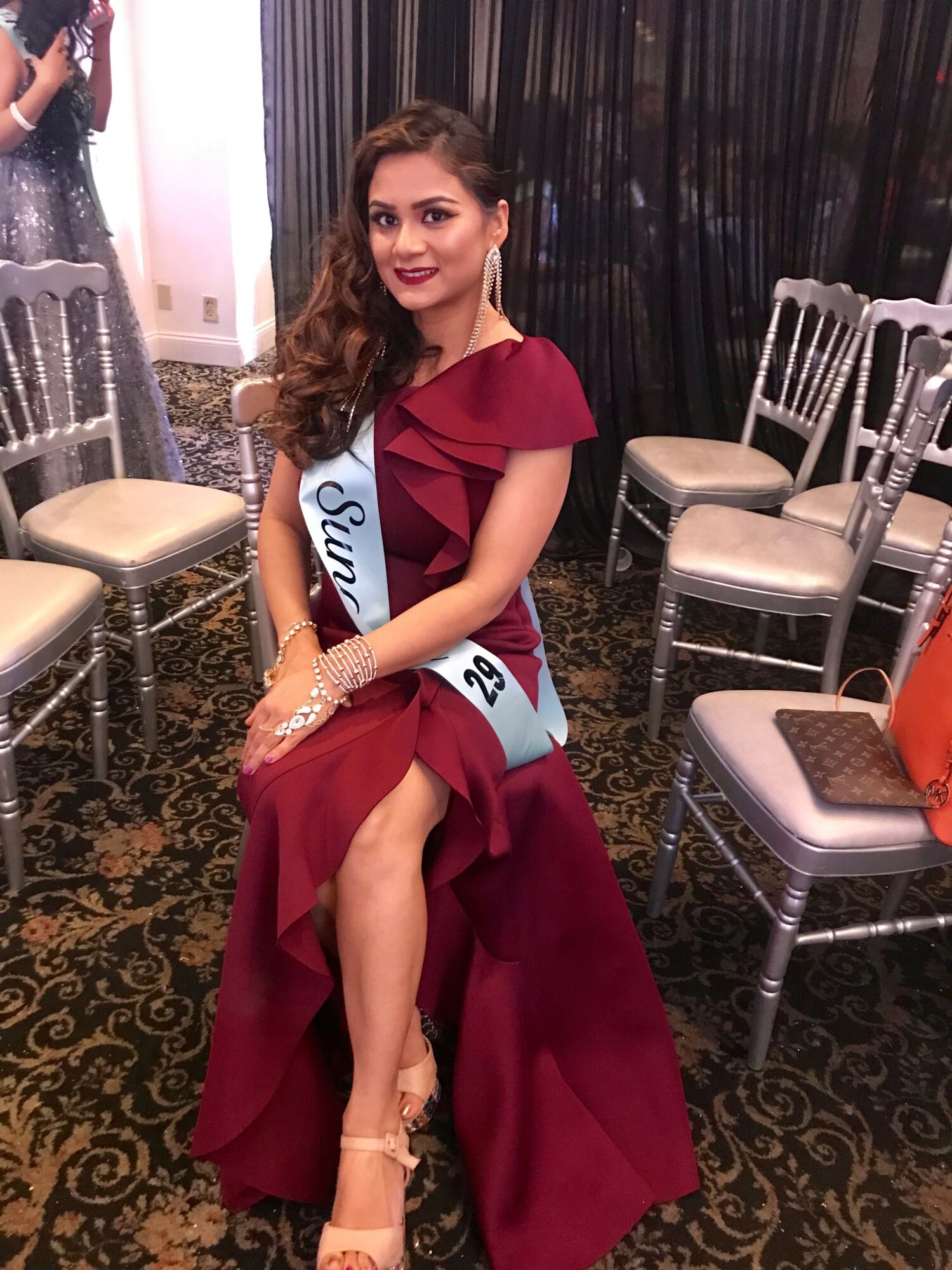 Woman in a red gown with a sash, sitting on a chair in a decorated room, smiling at the camera.