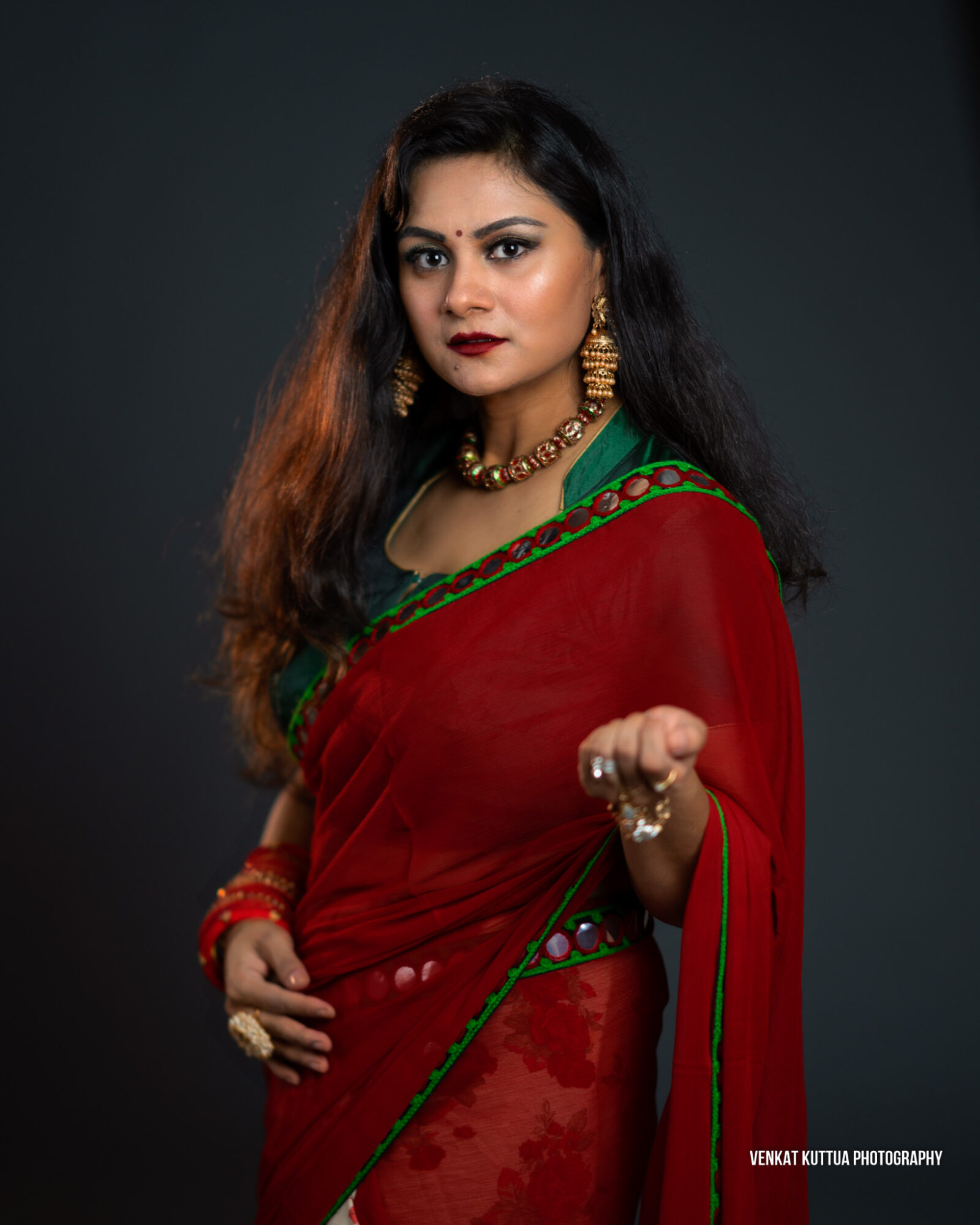 Woman in red and green traditional attire with jewelry, posing against a dark background.