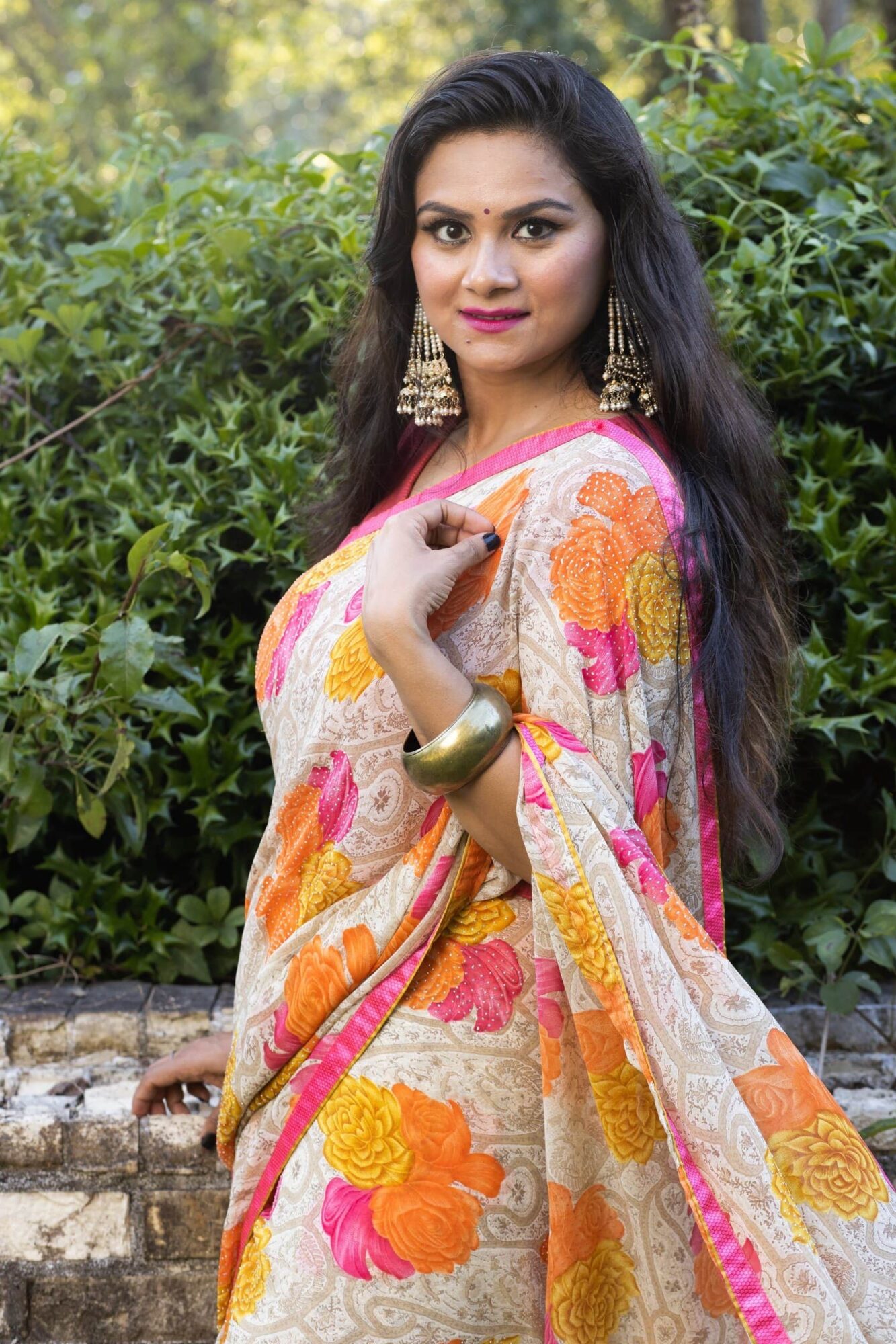 Woman in colorful saree with floral pattern, standing outdoors with greenery background, wearing earrings and bangles.
