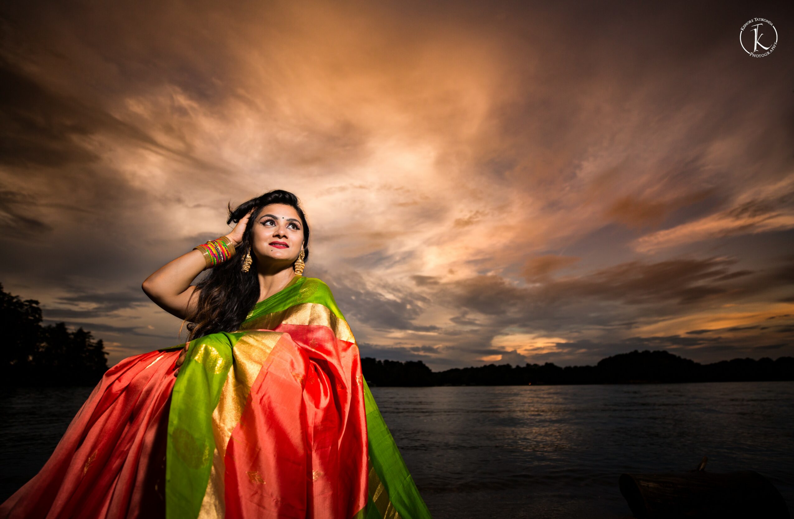 Woman in colorful traditional dress standing by water with dramatic sunset sky, clouds, and distant land in background.