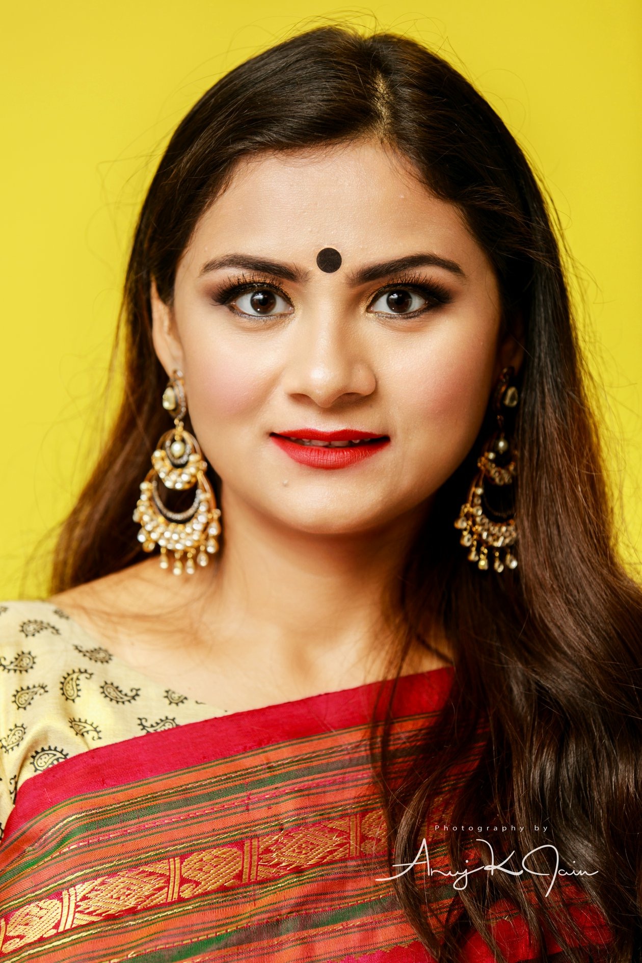 Young woman with long dark hair, wearing traditional earrings and saree, smiling against yellow background.