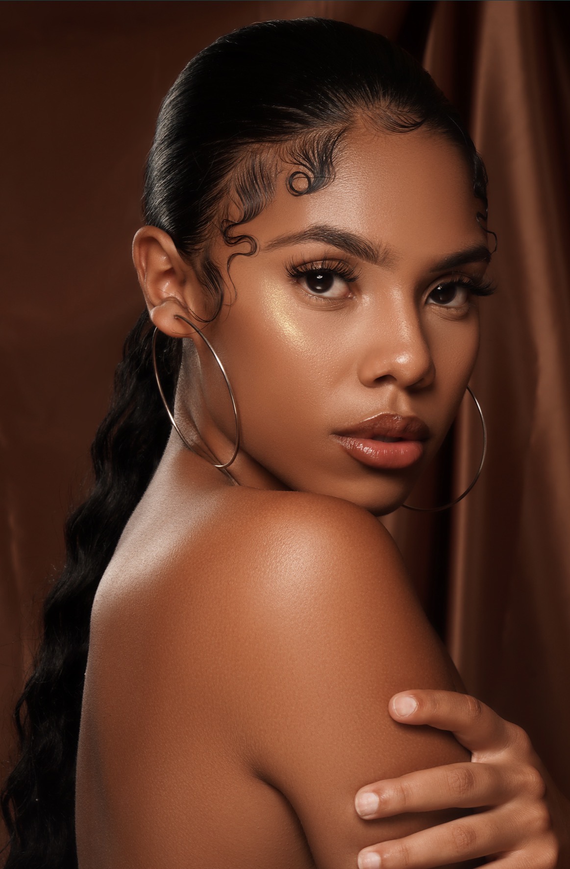 Close-up of a woman with styled hair, hoop earrings, and natural makeup, looking at the camera with a neutral expression.