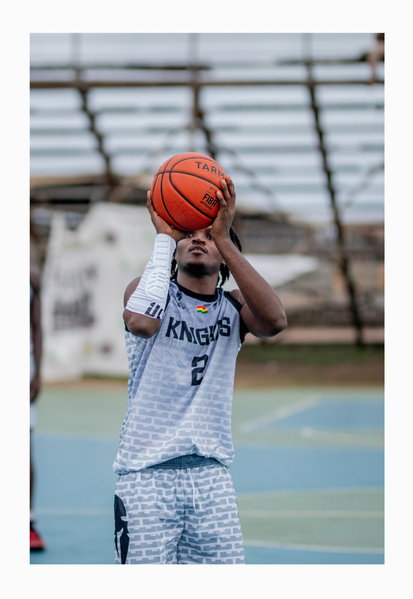 Child in basketball uniform shooting a basketball on an outdoor court, with bleachers in the background.