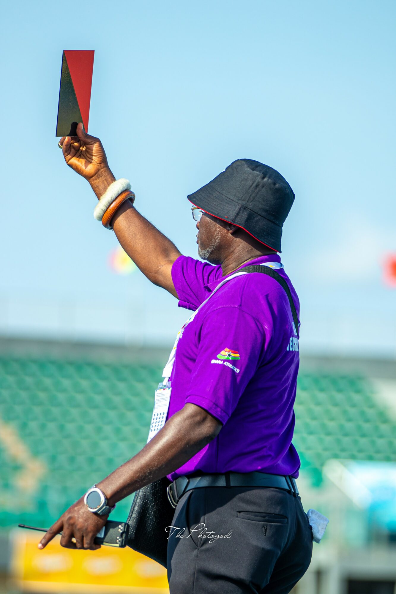 A man in a purple shirt and black hat holds a red and black flag, standing outdoors with a blue sky.