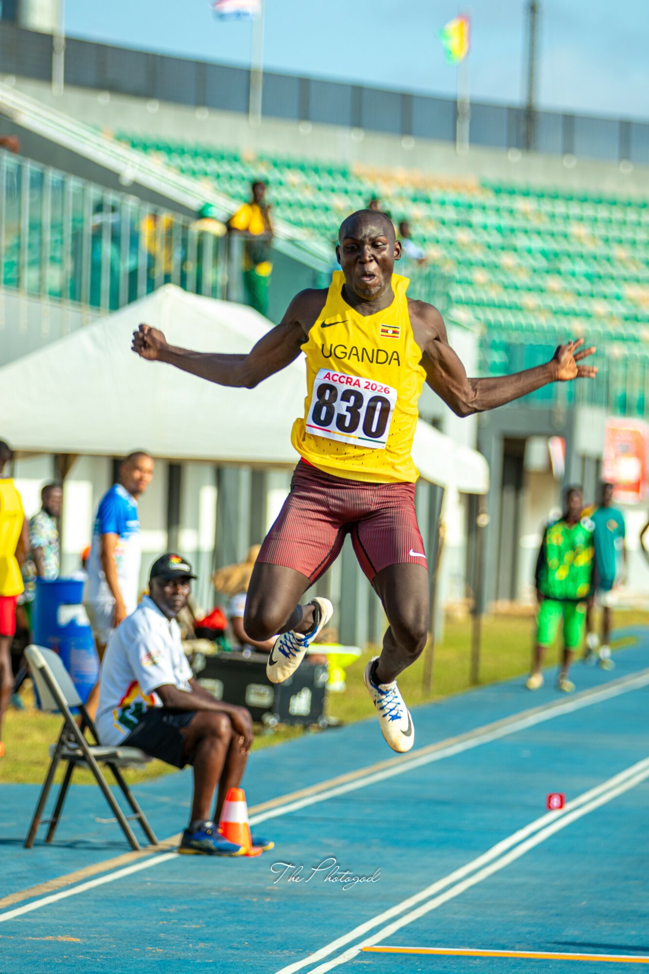 Male athlete in yellow and maroon uniform jumps on a blue track during a race, with stadium seats in background.