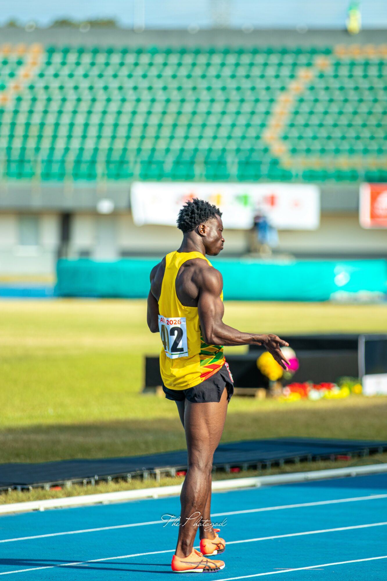 Male athlete standing on track holding a baton, wearing a yellow and black uniform, in a stadium.