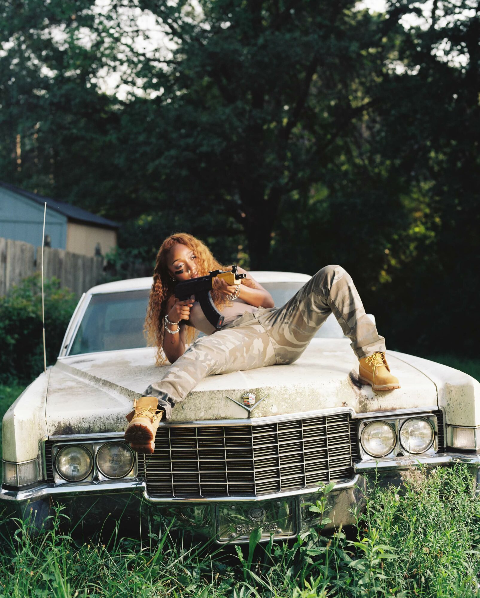 Person lying on the hood of a vintage car, holding a camera, outdoors with trees and grass around.
