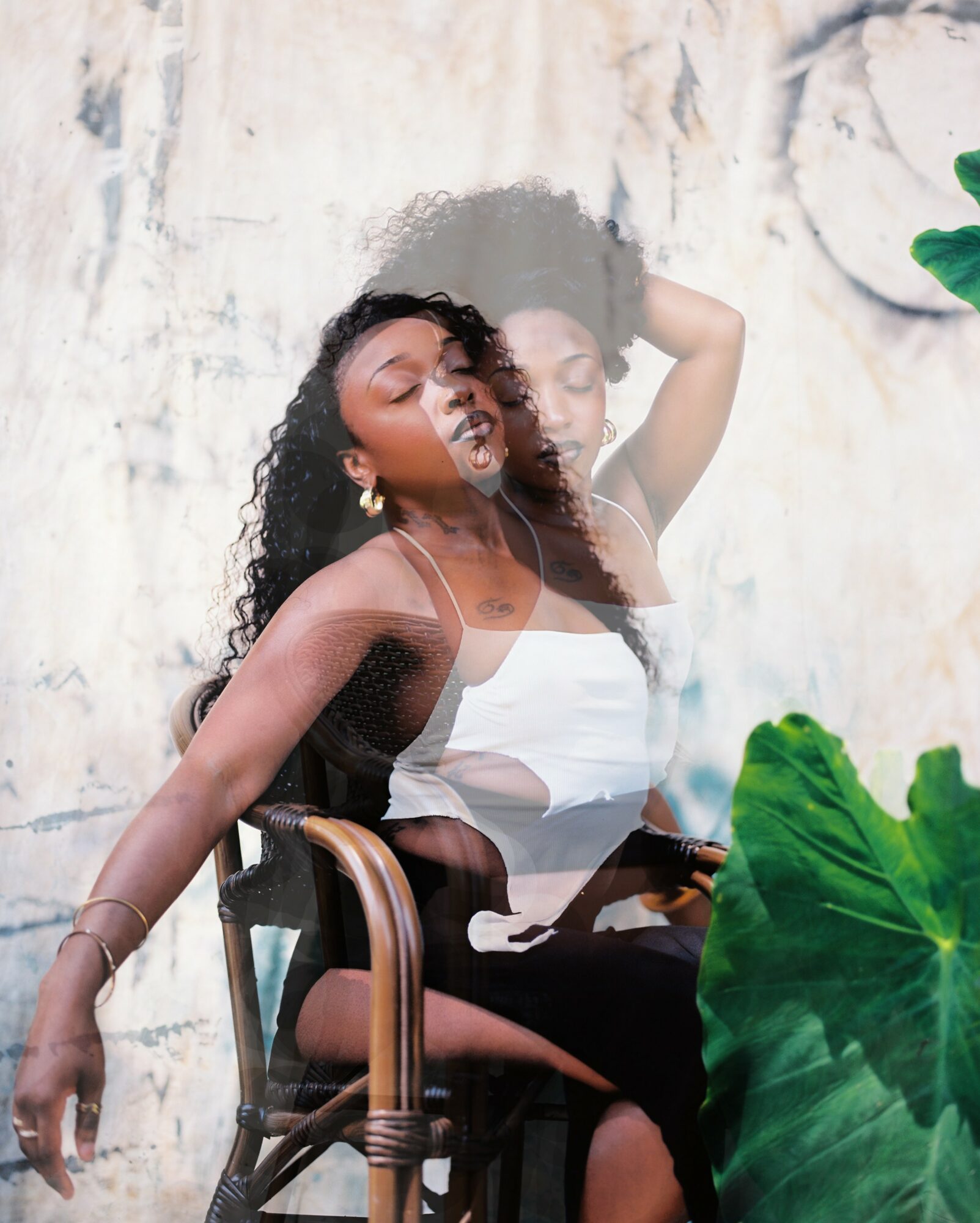 A woman with curly hair sitting on a chair, holding a phone, with a textured wall background and green leaves in the foreground.
