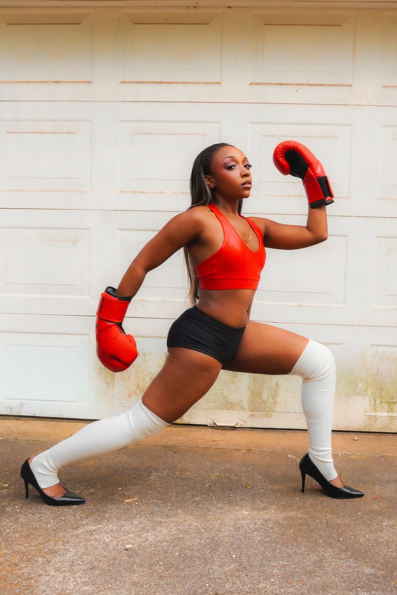 Woman in boxing gloves and athletic wear in a fighting stance outdoors, with a white garage door background.