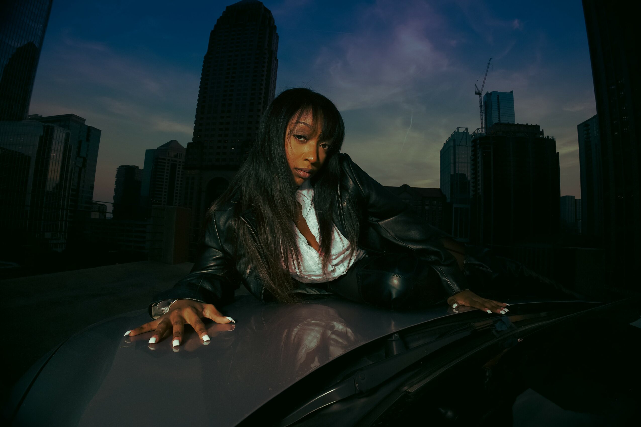 Woman with long dark hair leaning on a car hood in a city skyline at dusk.