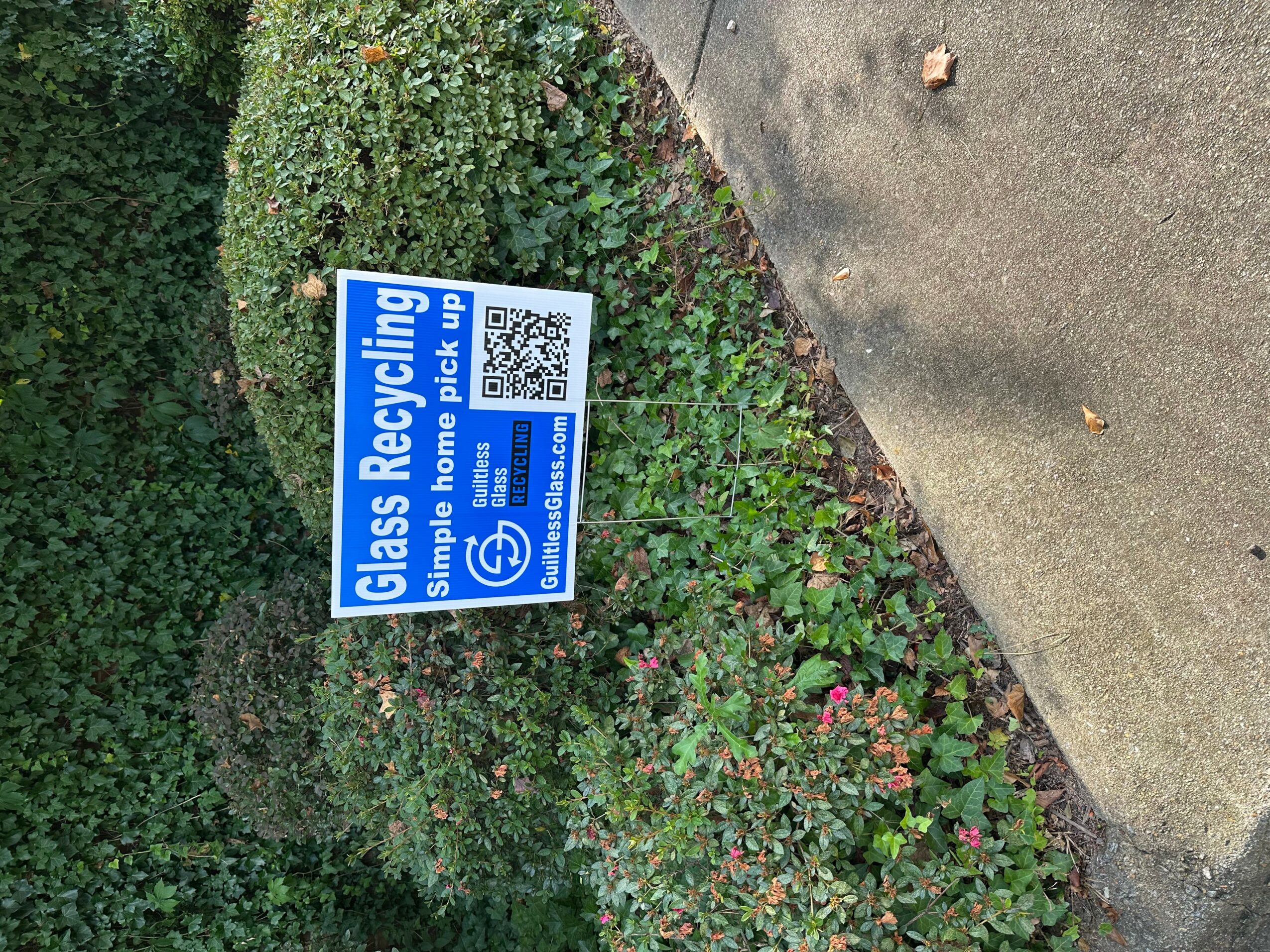 Blue sign indicating glass recycling with QR code, placed among bushes near a sidewalk.