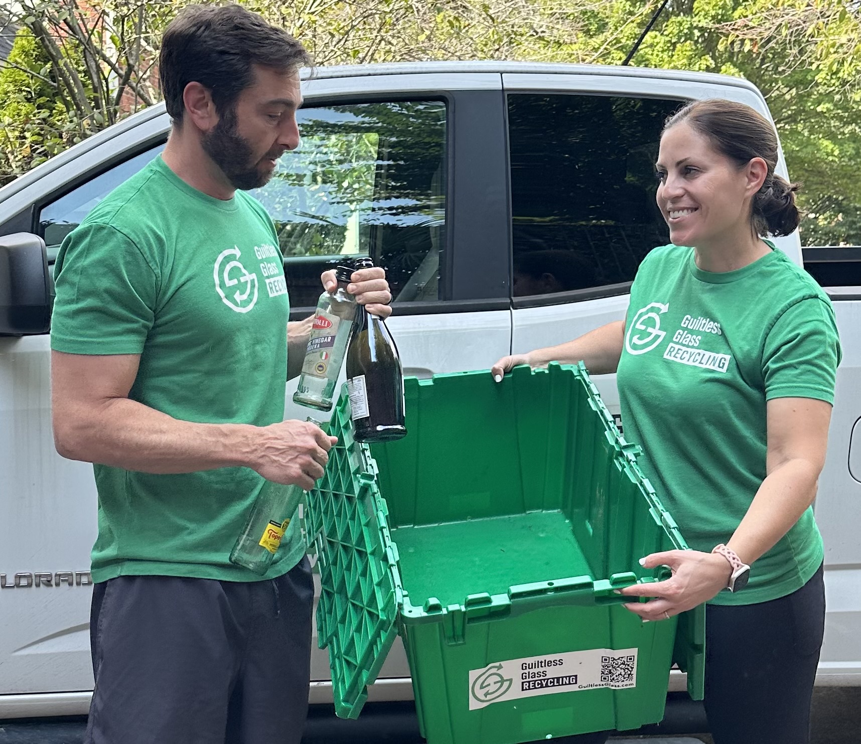 Two people in green shirts exchange bottles next to a vehicle, holding a green recycling bin outdoors.