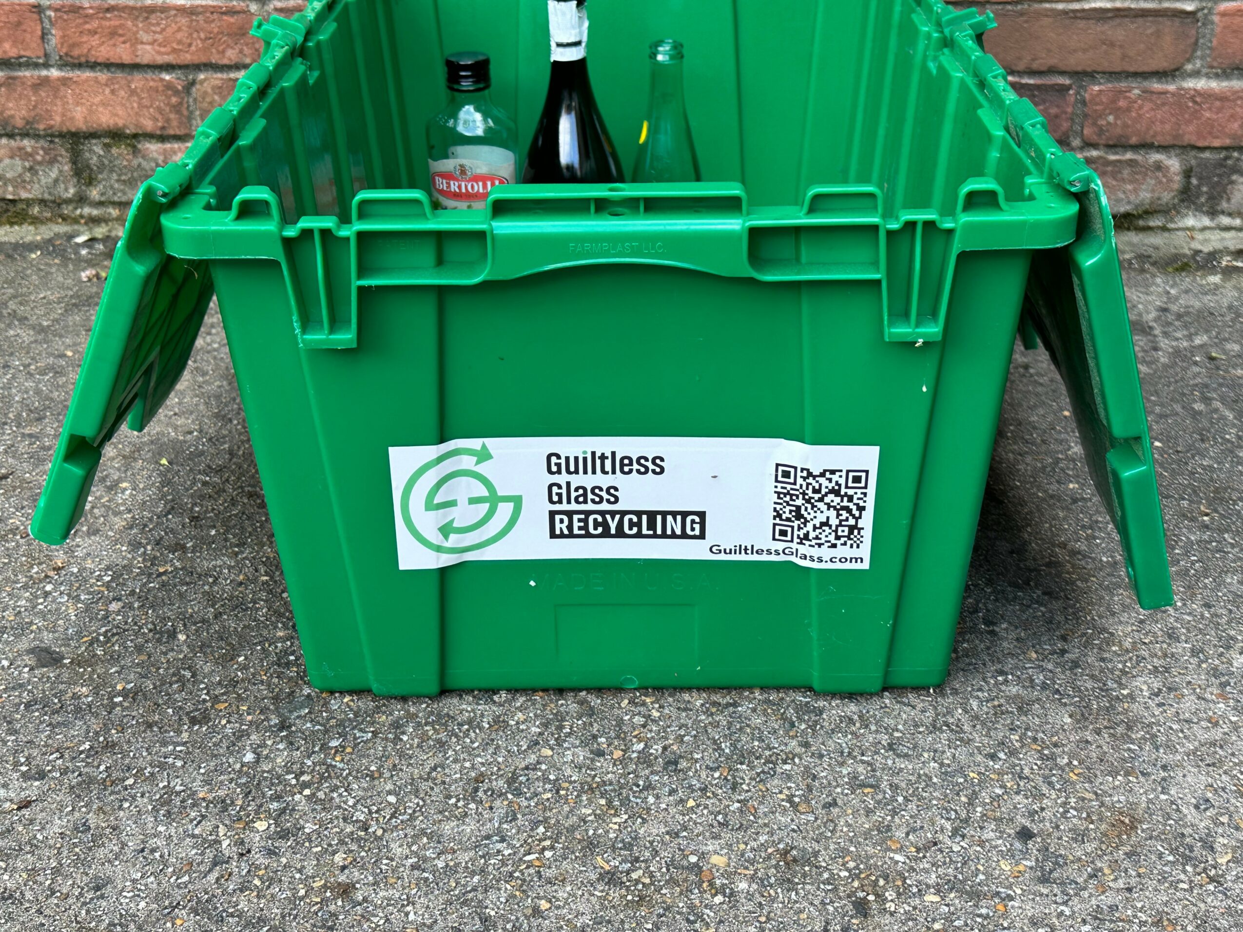 Green recycling bin with bottles inside, labeled for glass recycling, placed on pavement against a brick wall.