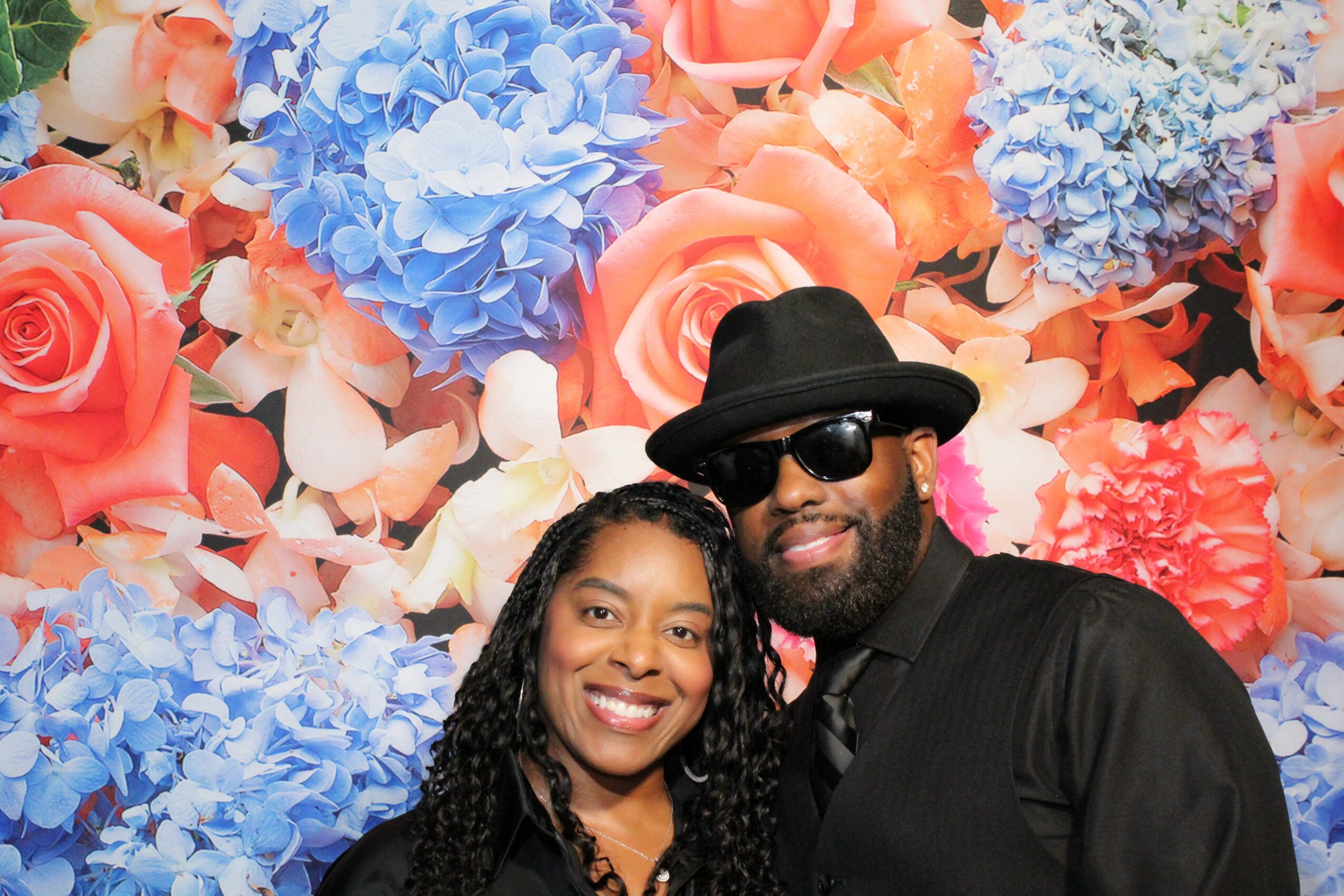 Two people smiling in front of a colorful floral background, one wearing sunglasses and a hat.