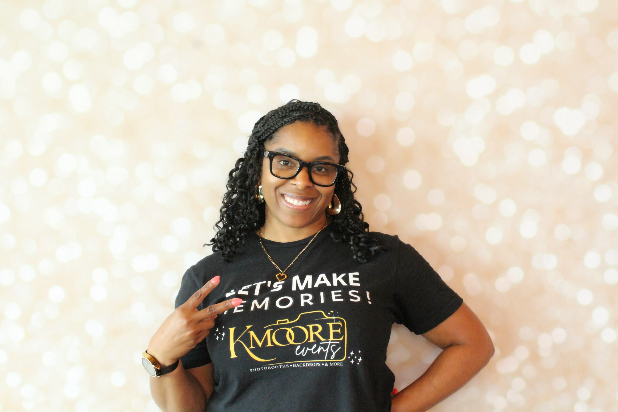 Woman with glasses smiling, wearing a black T-shirt with yellow text, making a peace sign gesture, standing against a light, bokeh background.