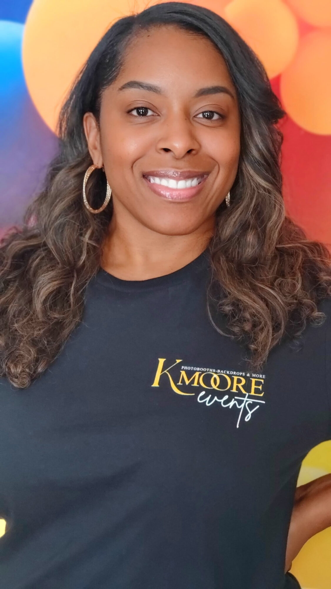 Woman with long wavy hair smiling, wearing a black shirt with yellow text, colorful background with balloons