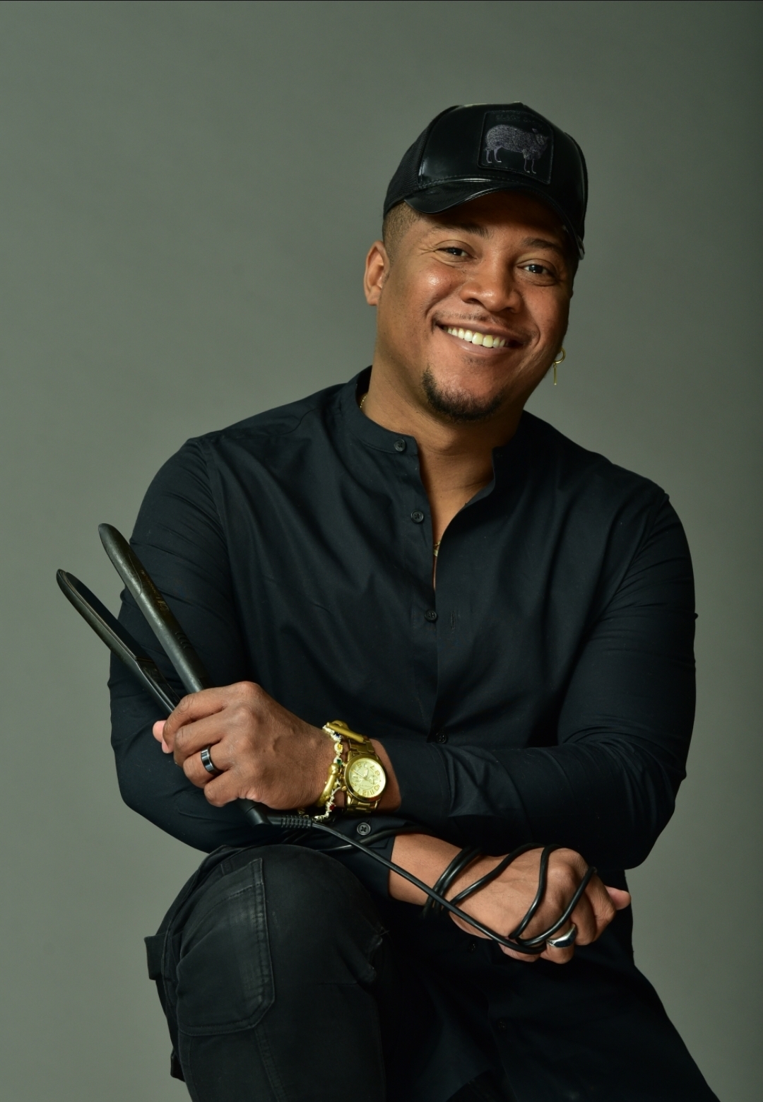 Smiling man wearing a black cap and shirt, holding a tablet and a phone, with a neutral background.