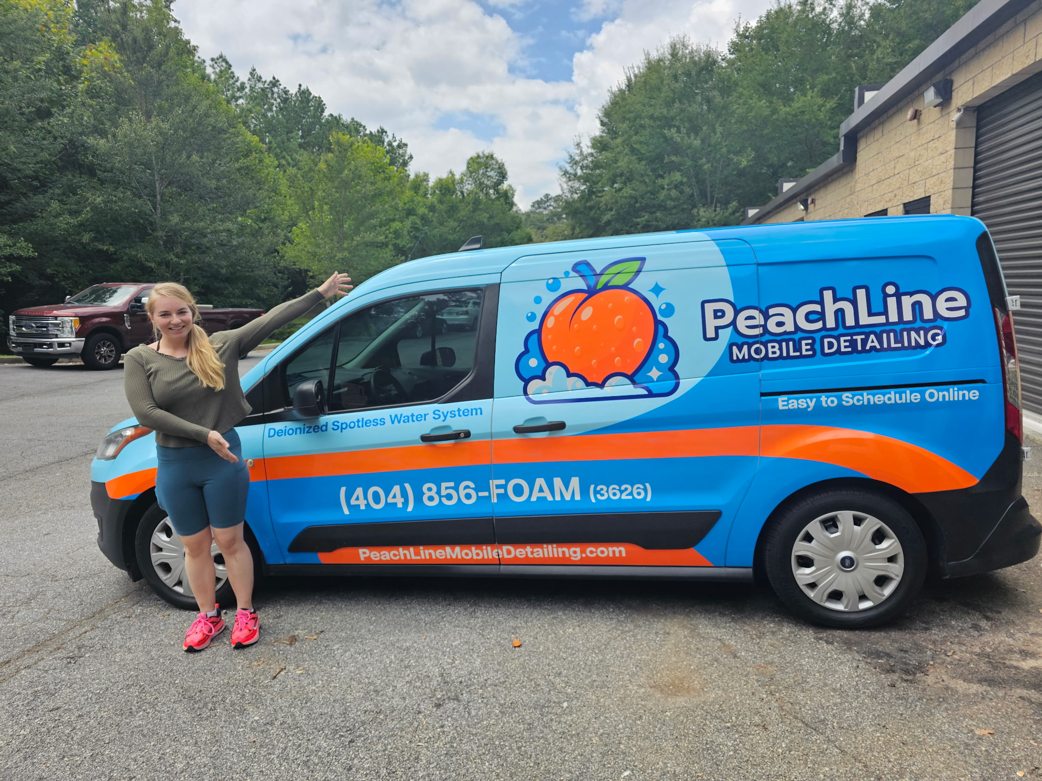 Woman standing next to a PeachLine mobile detailing van, pointing at the vehicle, parked outdoors with trees and a building in the background.