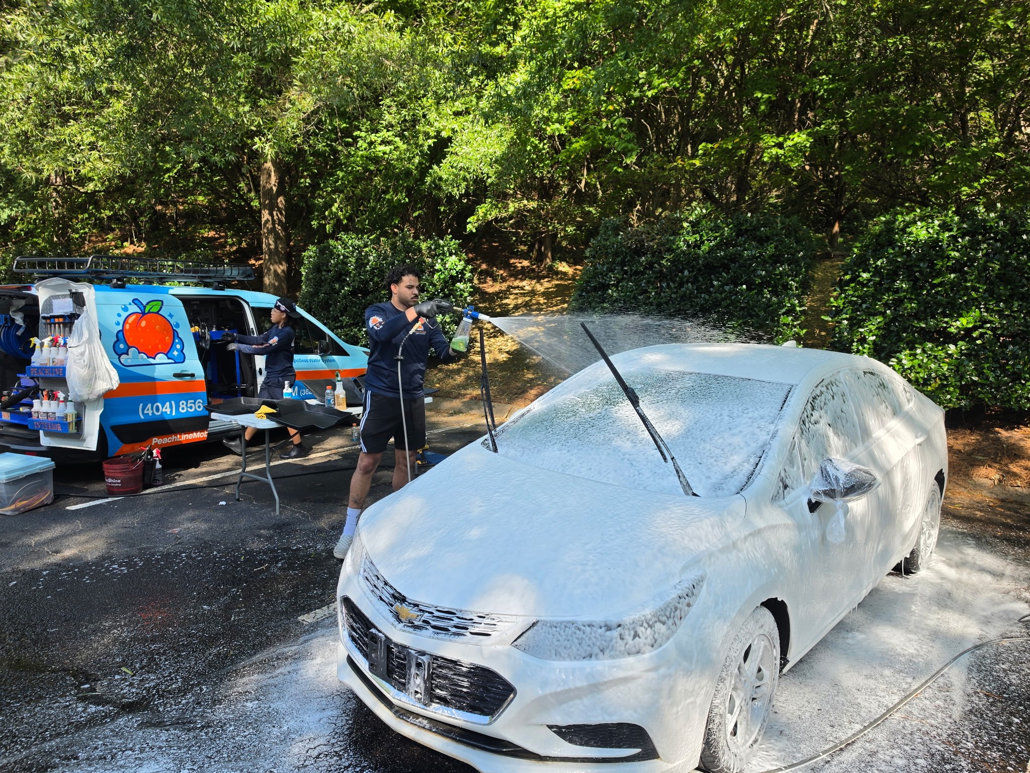 Person washing a white car with water spray outdoors, trees in background, police vehicle nearby, sunny day.