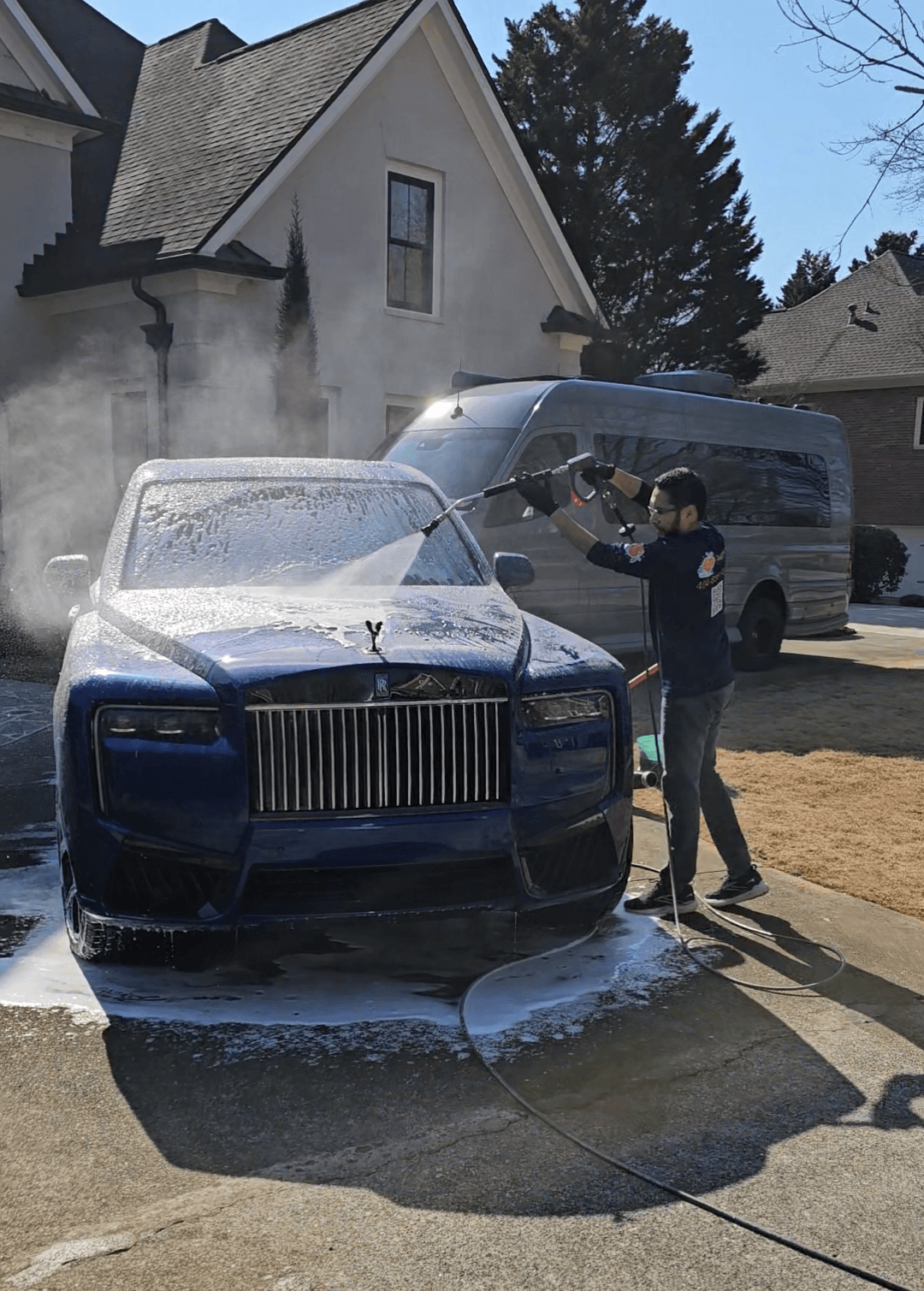 Person washing a dark-colored car with foam on the hood, driveway, house, and trees in background.