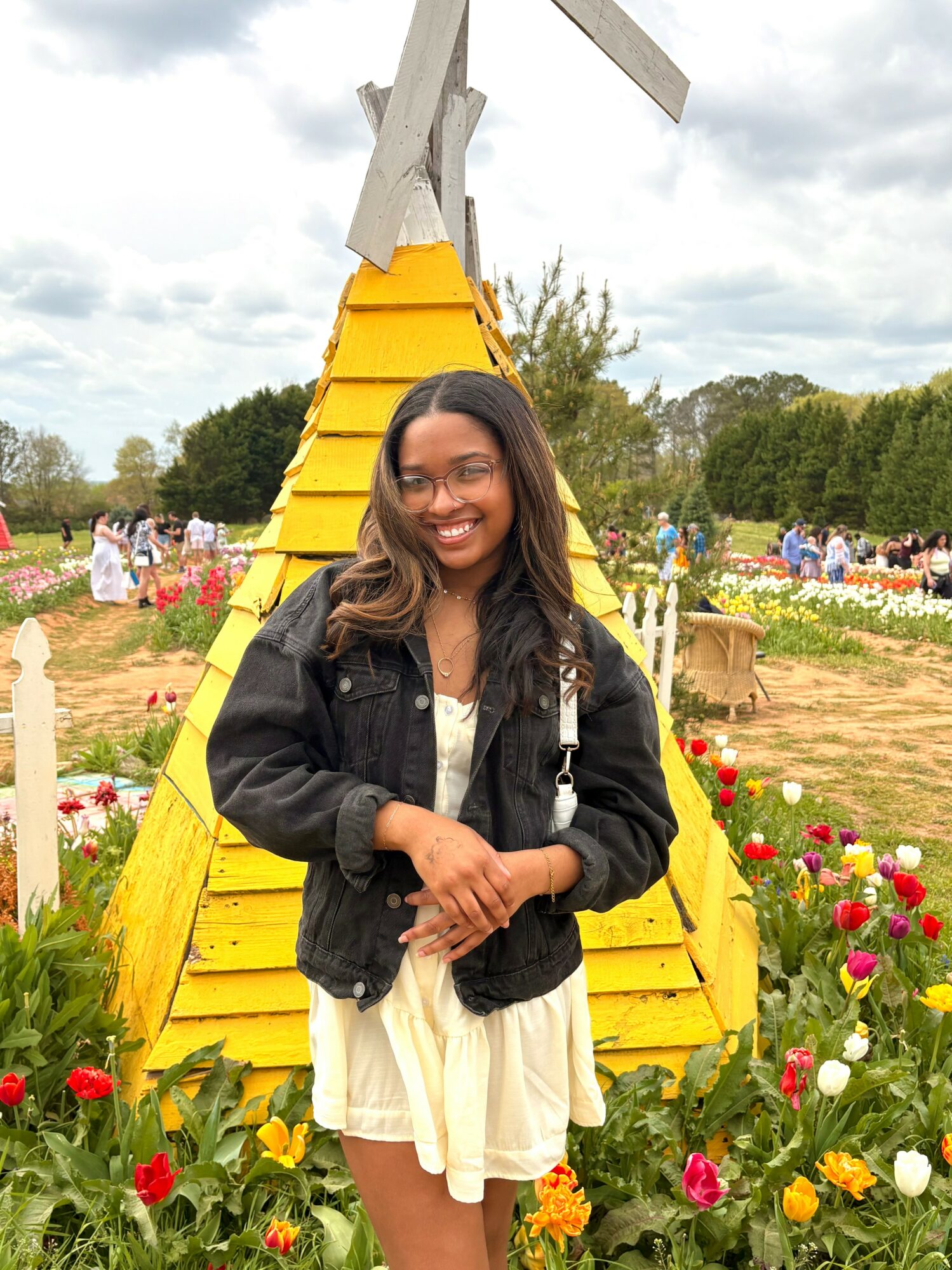 Smiling girl in a white dress and black jacket stands in front of a yellow wooden structure with flowers and a cloudy sky.