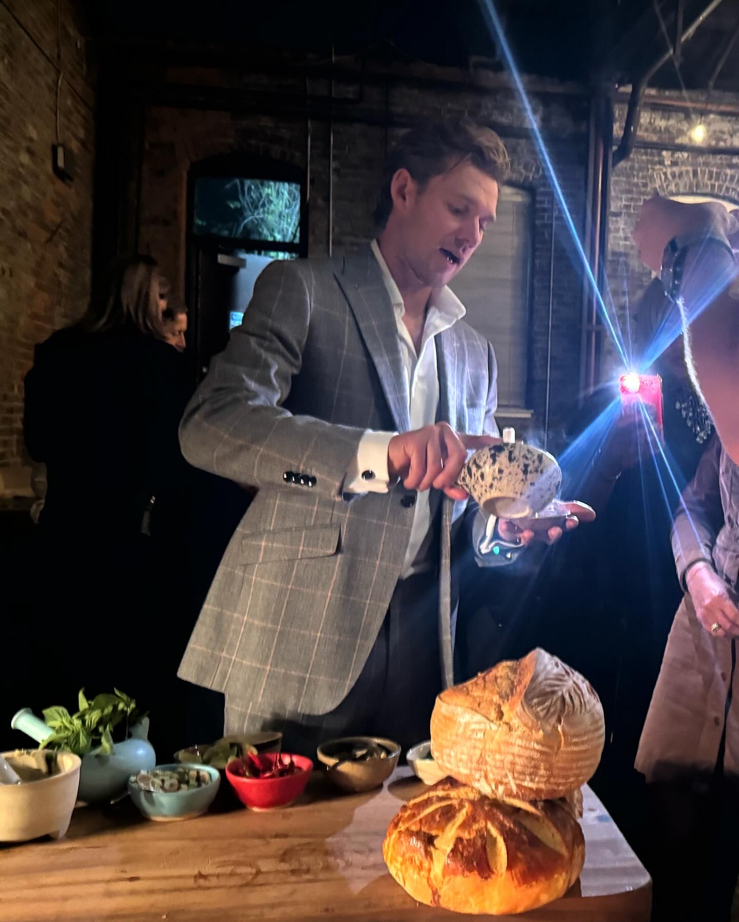 Man in gray suit pours drink into glass at bar with bread and bowls on table, people in background, dim lighting.