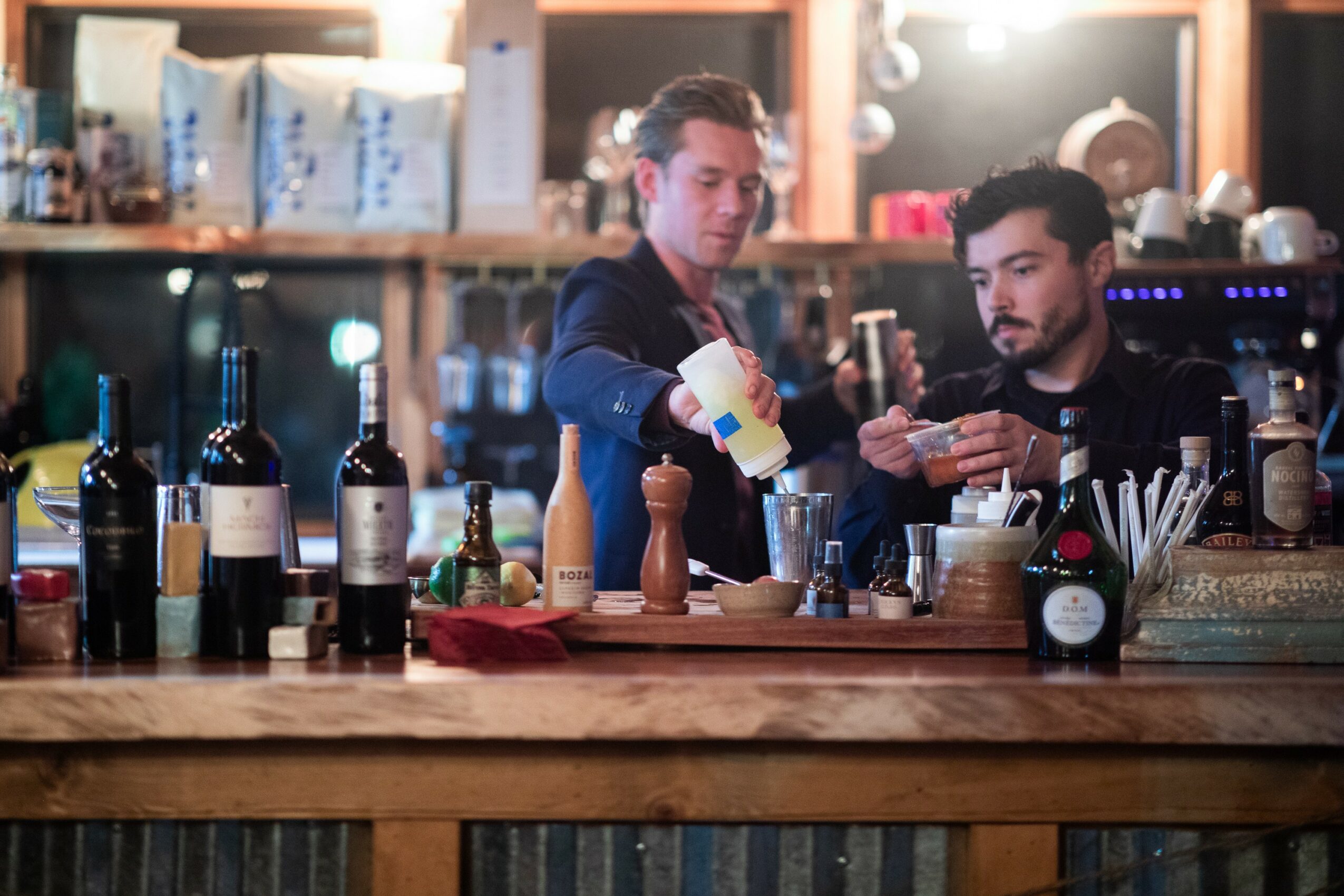 Two bartenders prepare drinks behind a bar with bottles and bar tools, in a dimly lit setting.