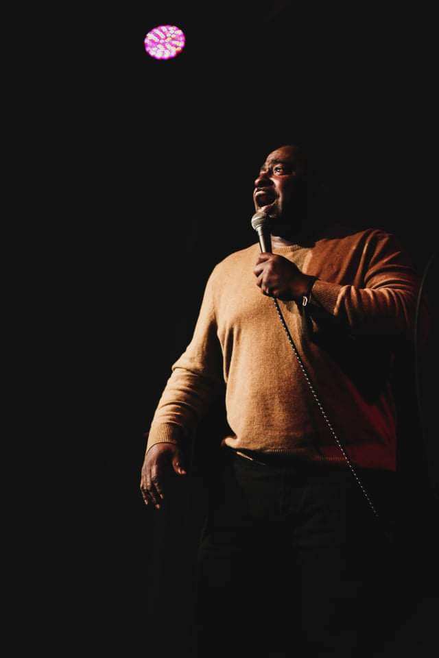 Person holding microphone on stage with a dark background and a pink light above, wearing a brown sweater.