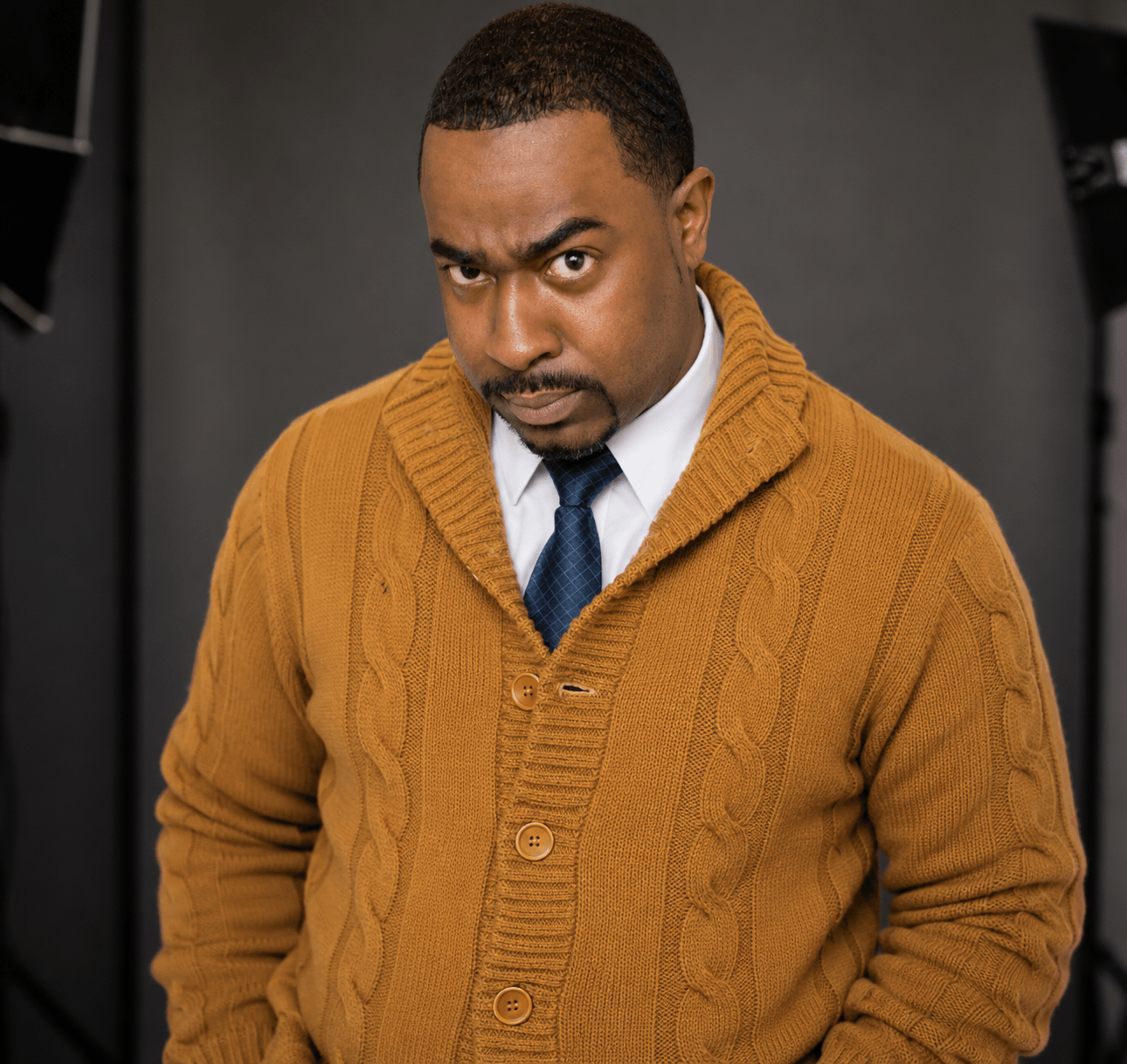 Man with serious expression wearing a mustard-colored sweater and white shirt, looking directly at the camera.