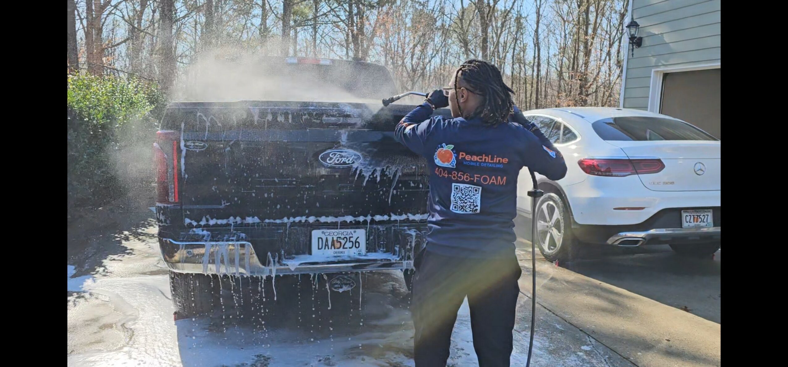 Person washing a black pickup truck with soap and water outdoors, other cars and trees in background.