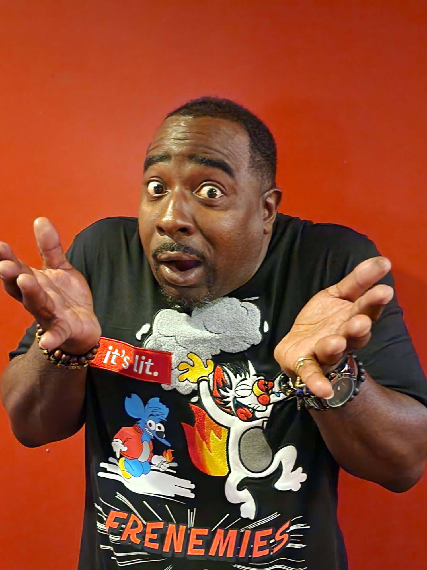 Man with surprised expression, hands raised, wearing a black T-shirt with cartoon characters, against an orange background.