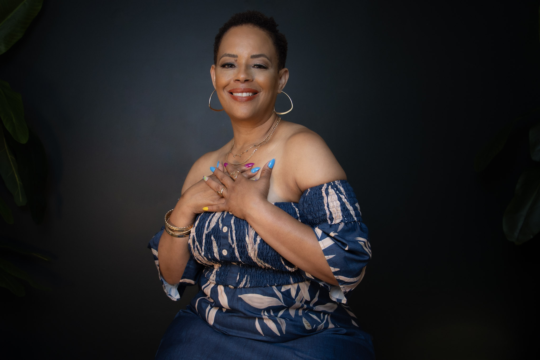Smiling woman with short hair, wearing hoop earrings and a patterned off-shoulder top, crosses her arms over her chest, against a dark background.