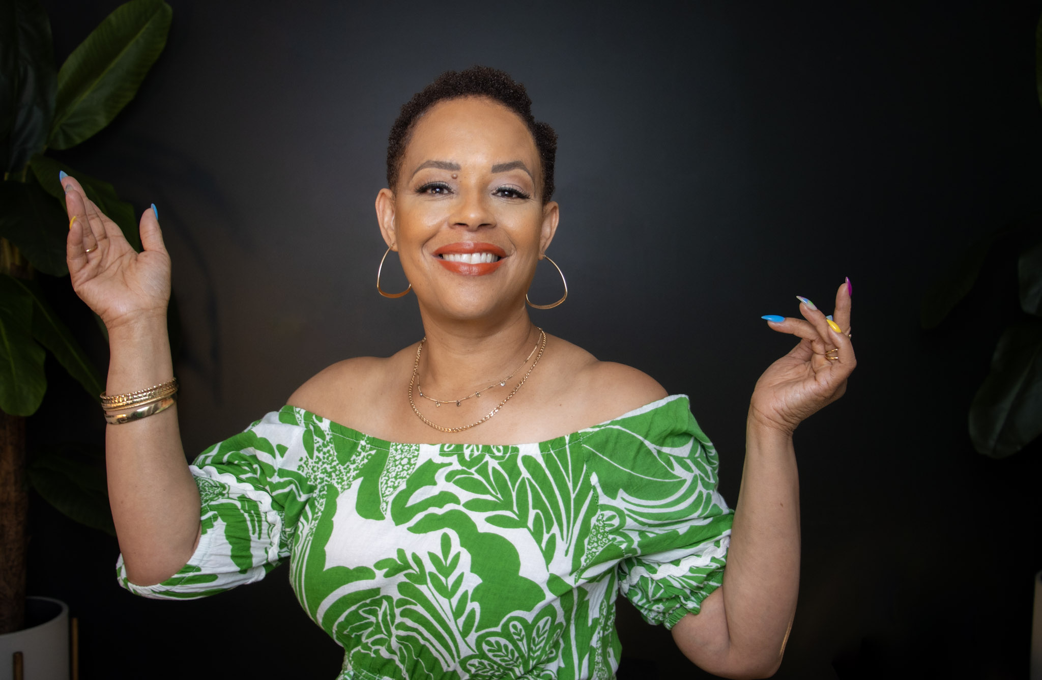 Smiling woman with short hair, wearing a green and white patterned off-shoulder top, raising her hands, against a dark background.